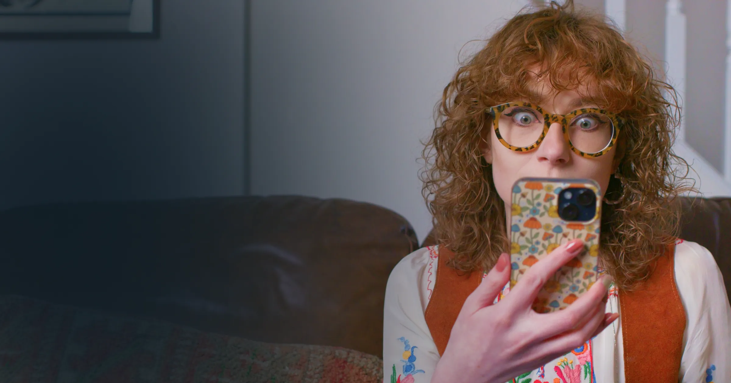 A surprised woman with curly hair and glasses holds a phone. Text reads.